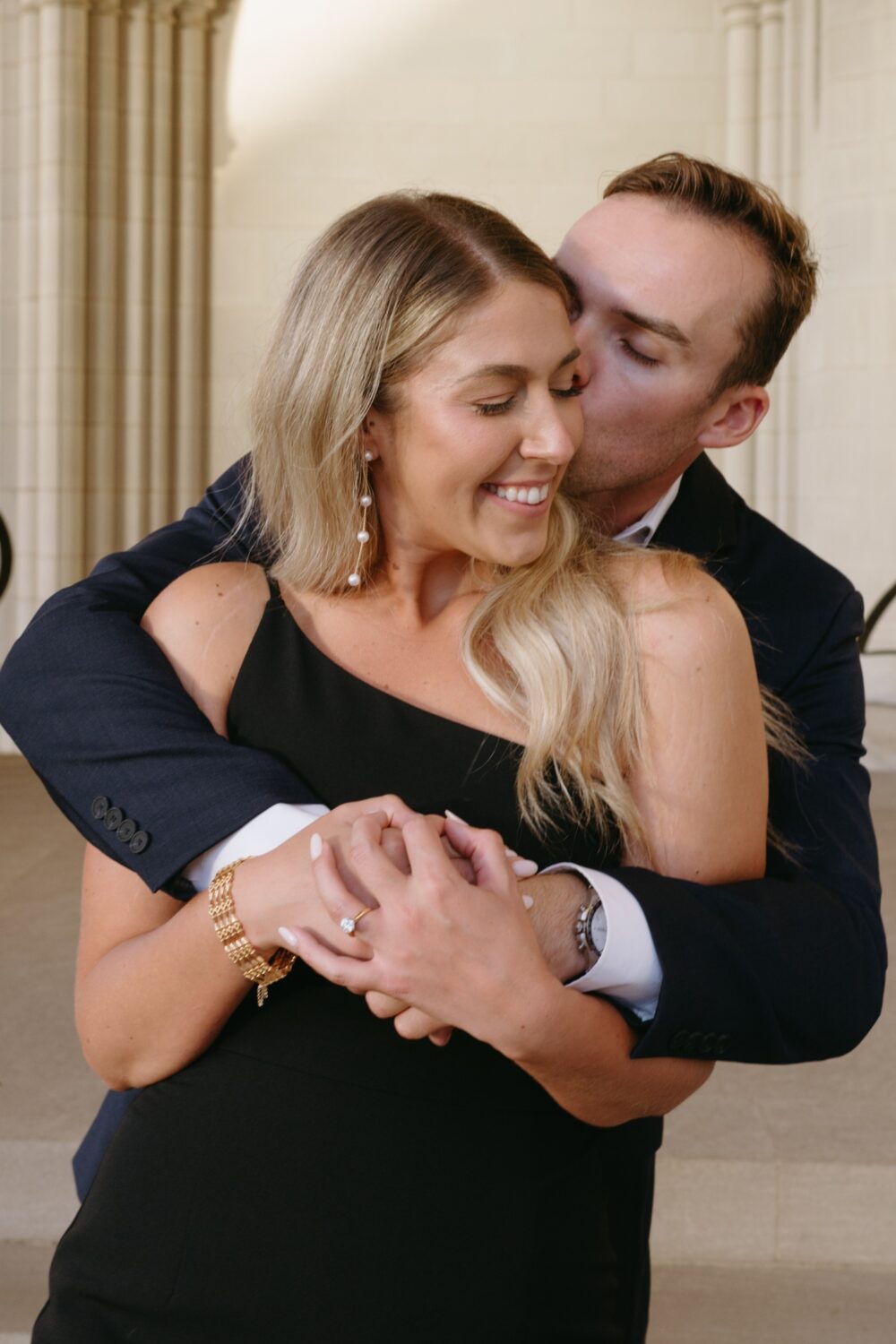 groom and bride to be hugging cheek kiss engagement ring details pearl earrings
