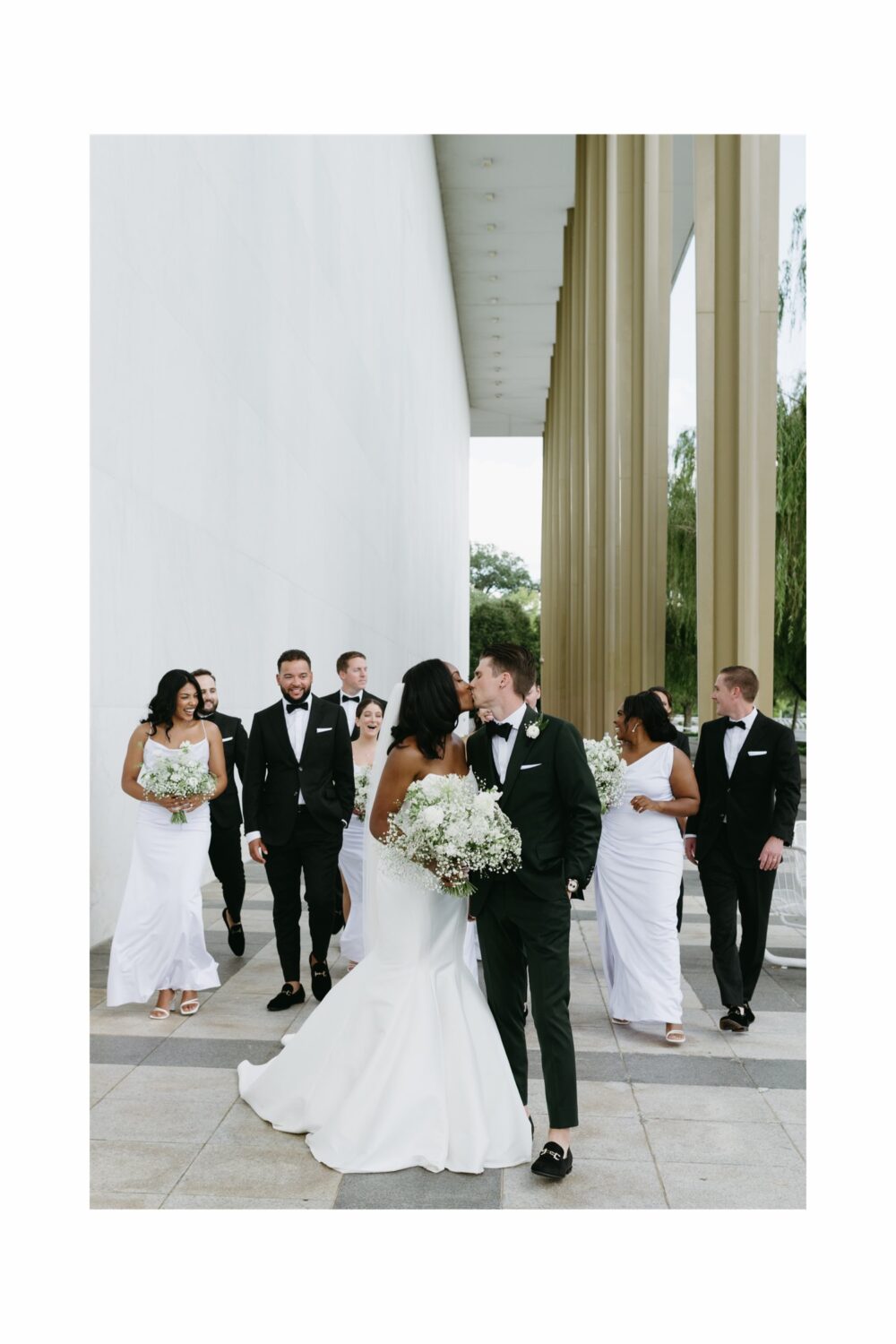 bride and groom kissing bridal party walking in background modern architecture 