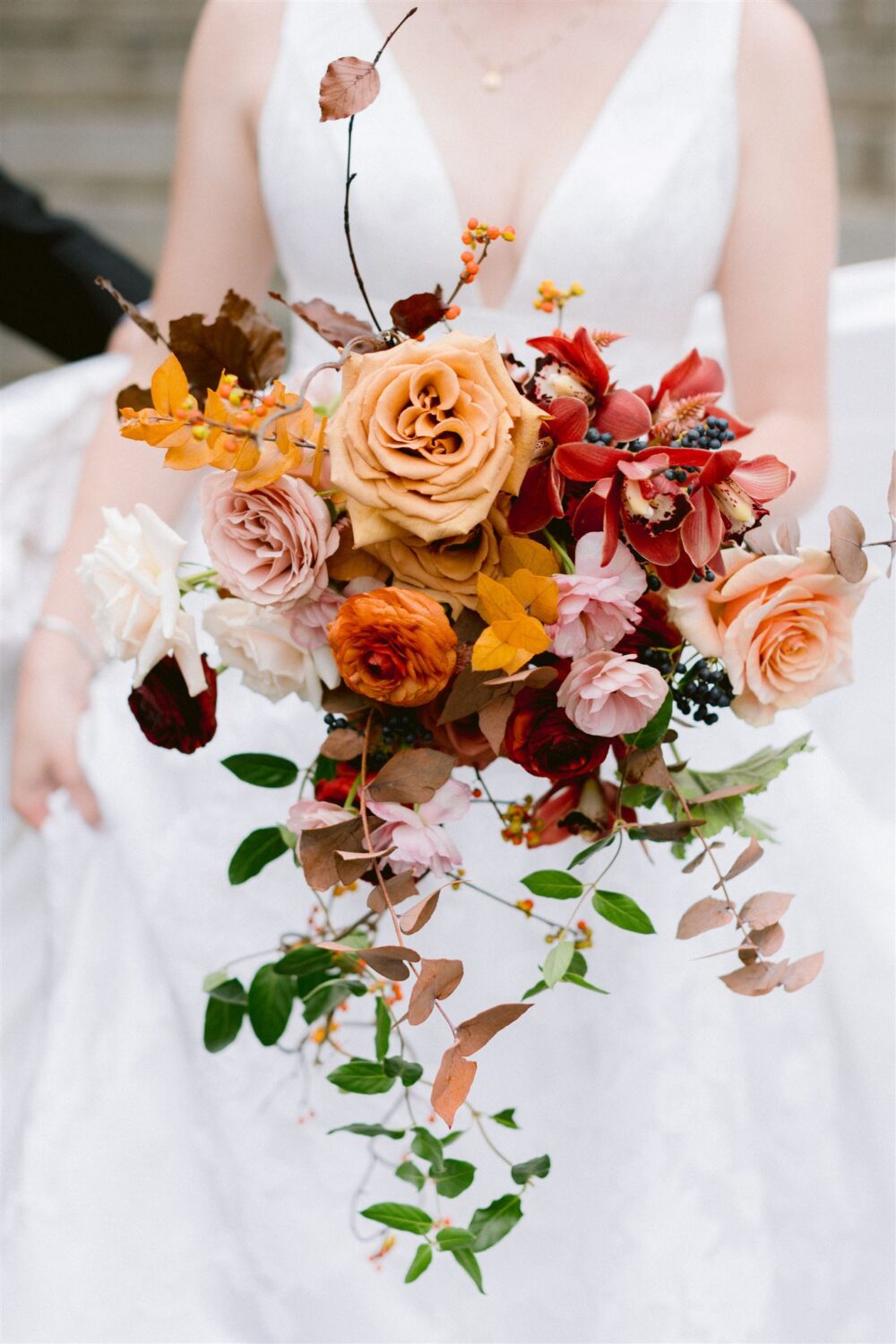 fall inspired wedding bouquet pink and cream roses red blooms berry accents against white of brides dress 